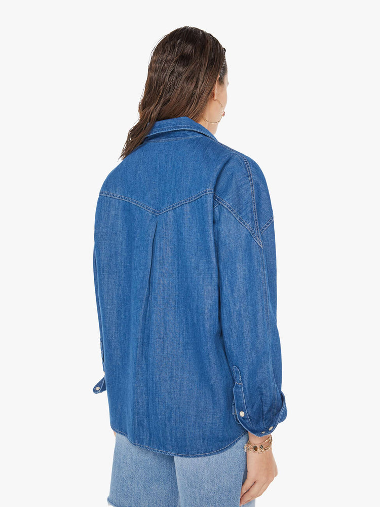 Back view of a woman wearing a dark blue wash denim shirt featuring an oversized fit, two large chest pockets, and white snap buttons, paired with a light blue wash short.