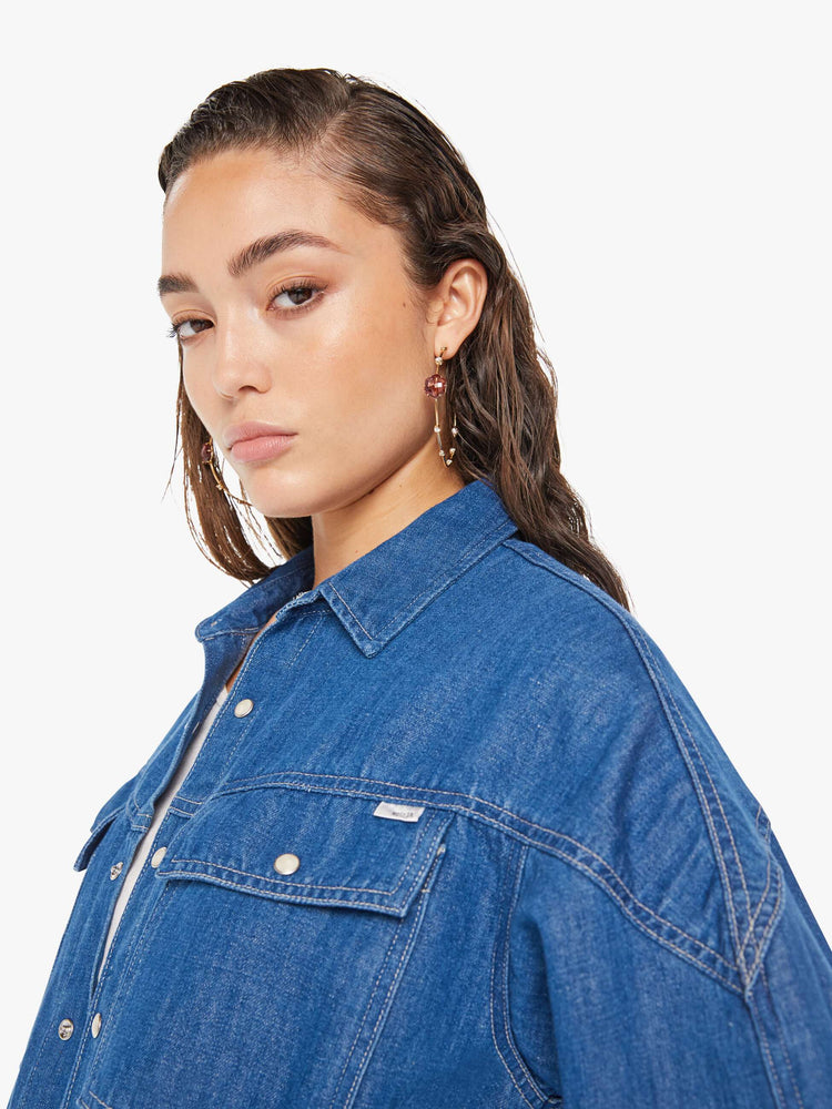 Side close up view of a woman wearing a dark blue wash denim shirt featuring an oversized fit, two large chest pockets, and white snap buttons.