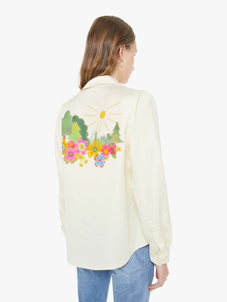 Back  view of a woman in an off white with colored flowers collared button-up with long gathered sleeves and a cropped, curved hem.