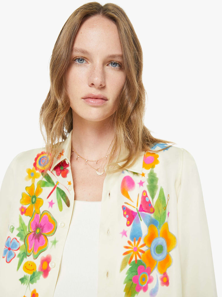 Close up  view of a woman in an off white with colored flowers collared button-up with long gathered sleeves and a cropped, curved hem.