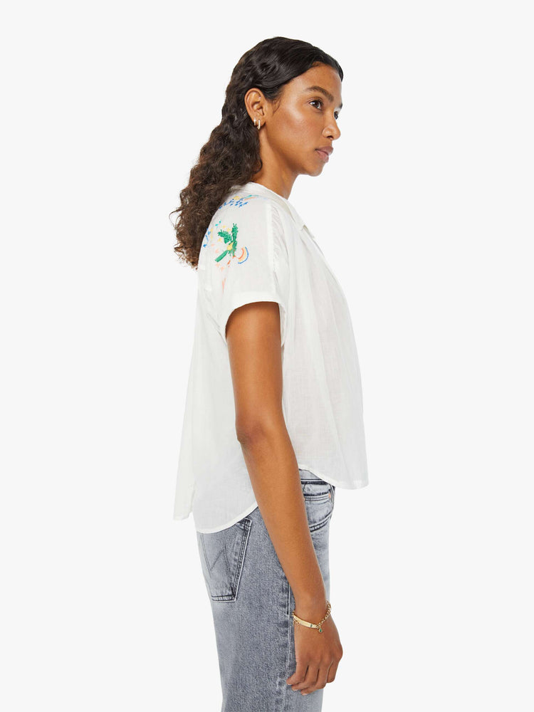 Side view of a woman in a short-sleeved button-up with a curved hem, gathered seams and embroidered tropical motifs across the back. Paired with grey jeans.