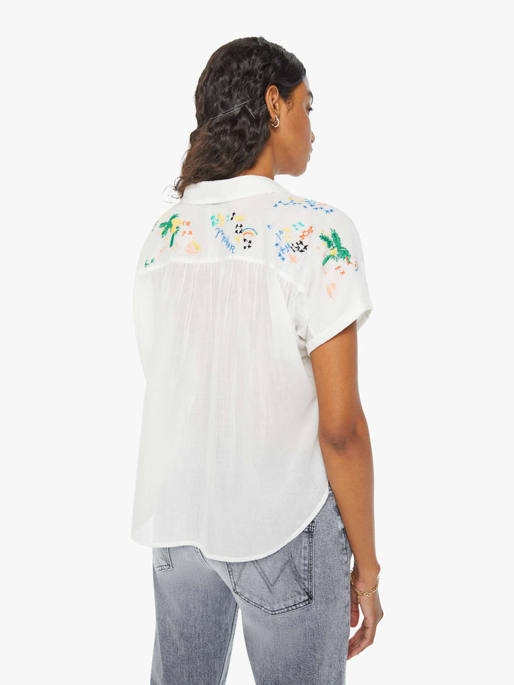 Back view of a woman in a short-sleeved button-up with a curved hem, gathered seams and embroidered tropical motifs across the back. Paired with grey jeans.
