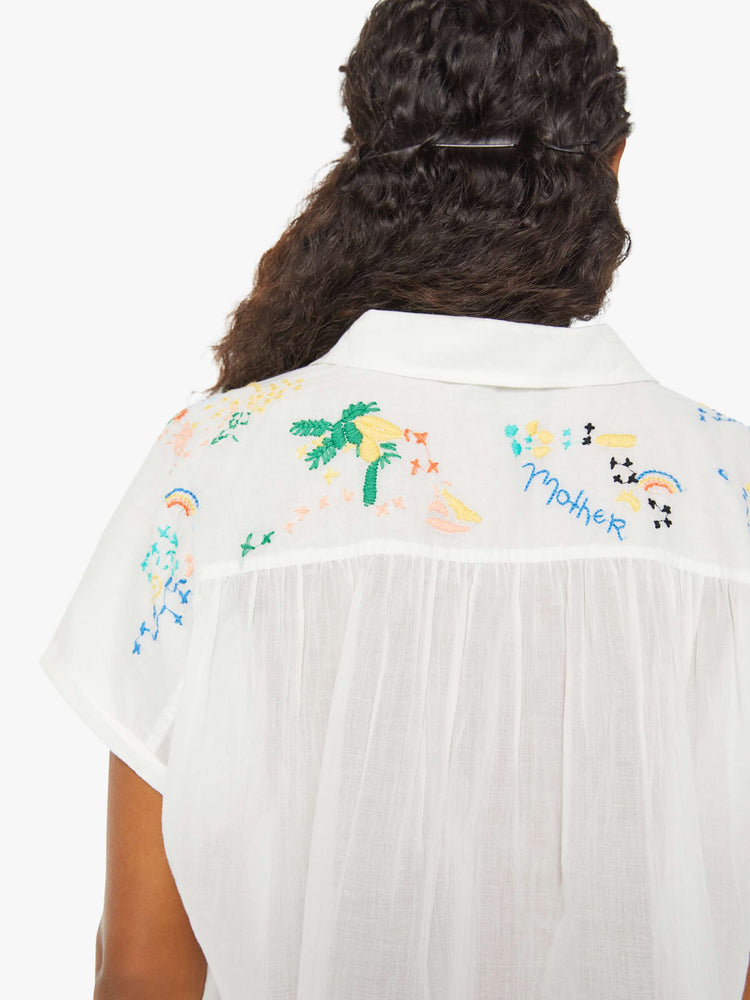 Detailed view of a woman in a short-sleeved button-up with a curved hem, gathered seams and embroidered tropical motifs across the back. 