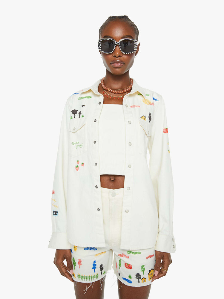 Front view of a woman in a white with embroidery throughout denim button-up with front patch pockets and a curved hem.