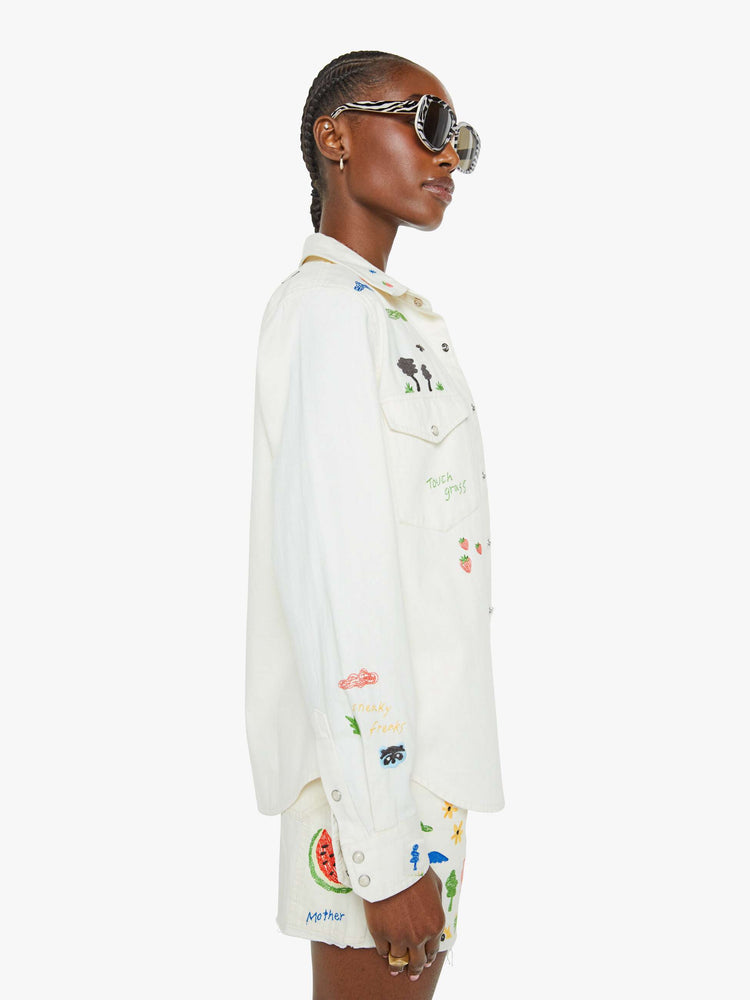 Side view of a woman in a white with embroidery throughout denim button-up with front patch pockets and a curved hem.