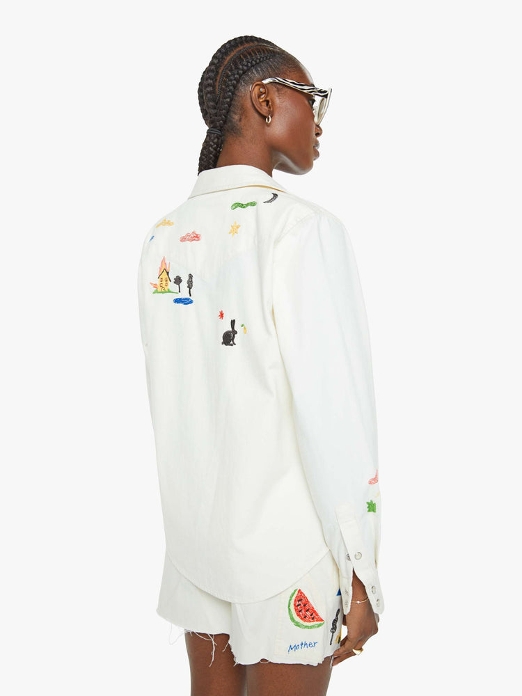 Back view of a woman in a white with embroidery throughout denim button-up with front patch pockets and a curved hem.