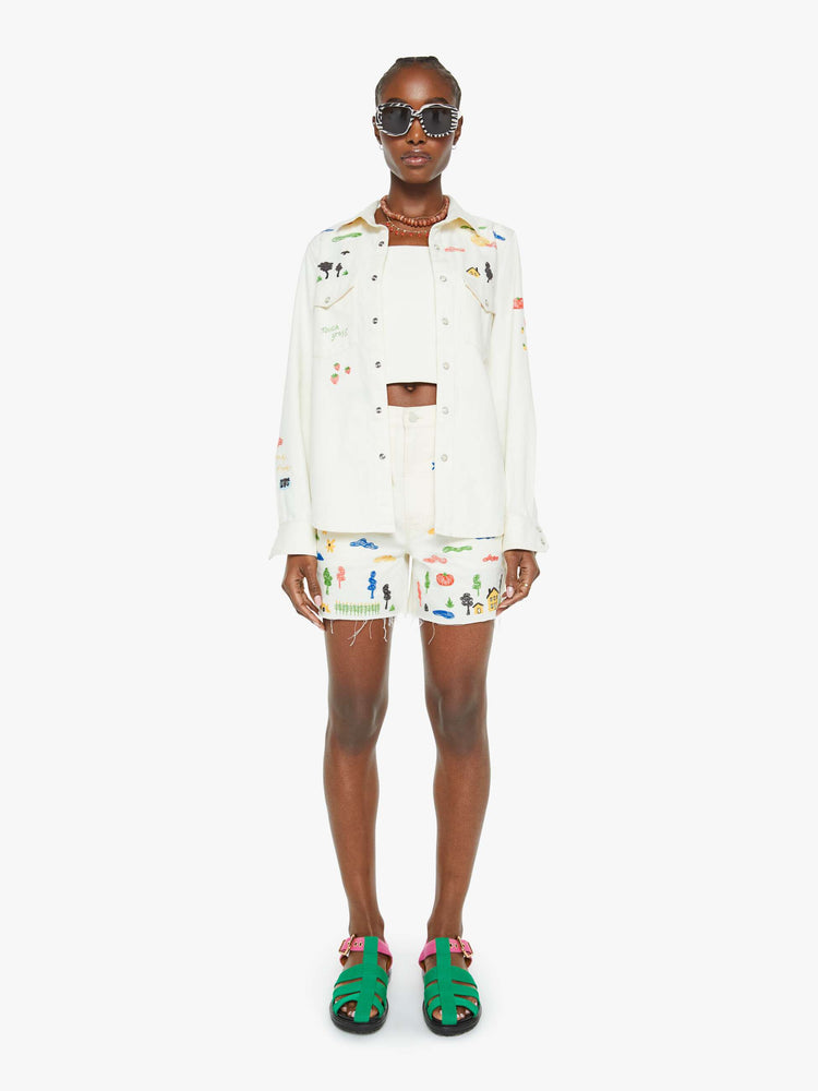 Full body view of a woman in a white with embroidery throughout denim button-up with front patch pockets and a curved hem.