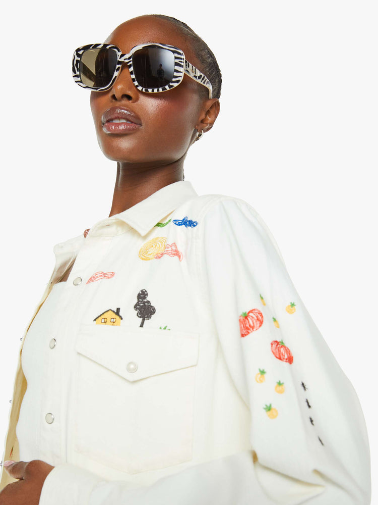 Close up view of a woman in a white with embroidery throughout denim button-up with front patch pockets and a curved hem.