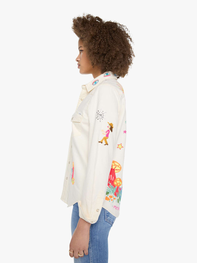 Left side view of a woman in denim button-up with front patch pockets and a curved hem creamy off-white with colorful embroidered mushrooms, stars and text throughout.