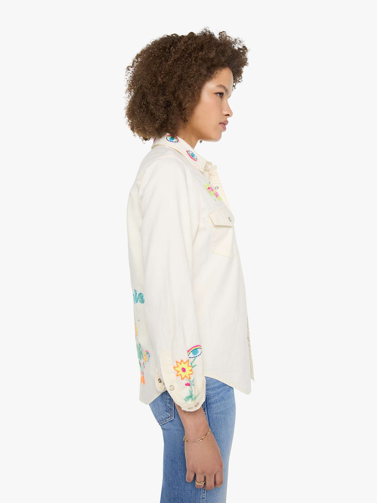 Side view of a woman in denim button-up with front patch pockets and a curved hem creamy off-white with colorful embroidered mushrooms, stars and text throughout.