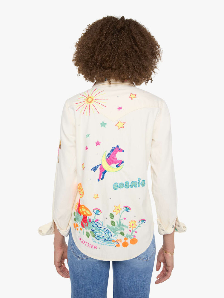 Back view of a woman in denim button-up with front patch pockets and a curved hem creamy off-white with colorful embroidered mushrooms, stars and text throughout.