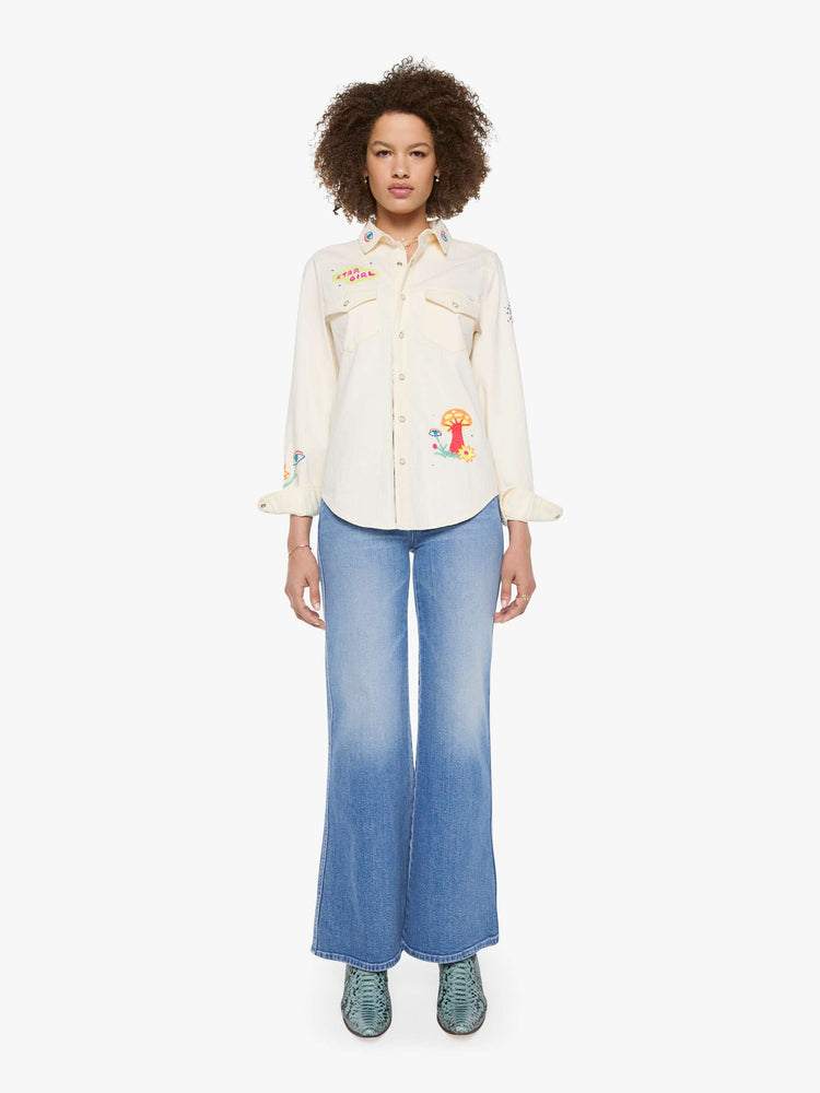 Full body view of a woman in denim button-up with front patch pockets and a curved hem creamy off-white with colorful embroidered mushrooms, stars and text throughout.