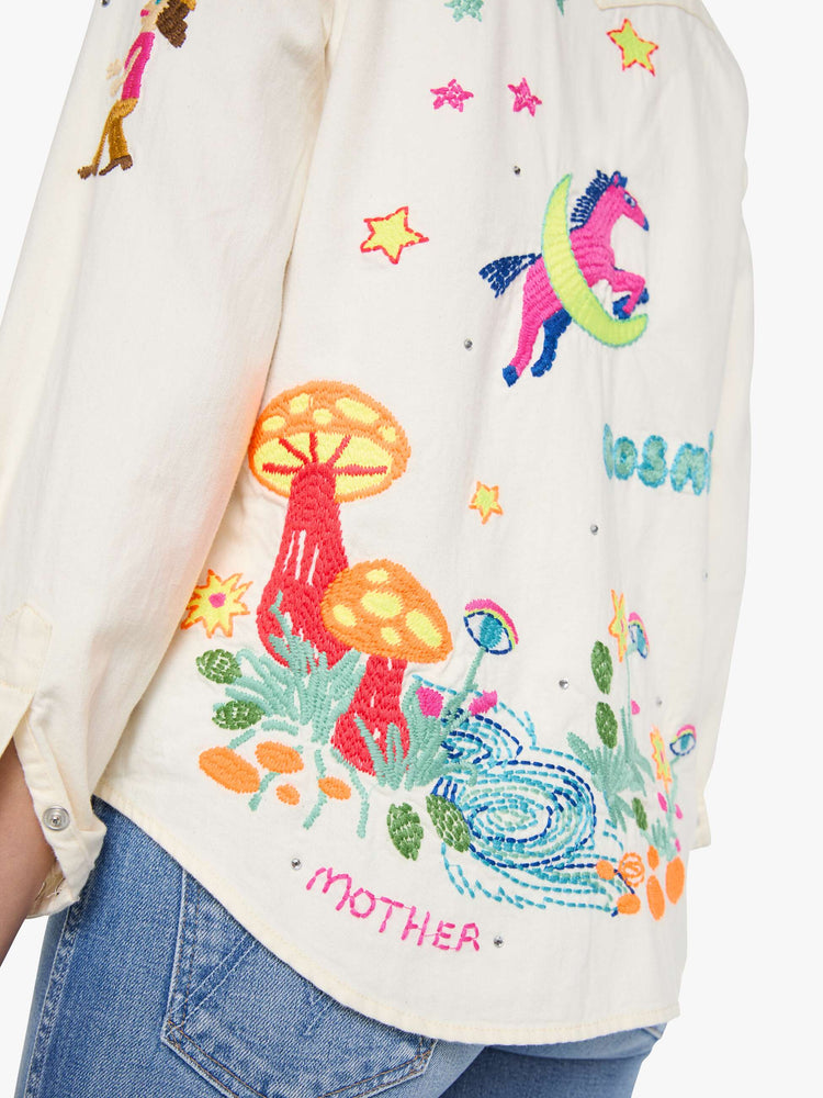 Detailed shot view of a woman in denim button-up with front patch pockets and a curved hem creamy off-white with colorful embroidered mushrooms, stars and text throughout.