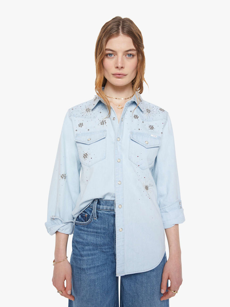 Front view of a woman in a faded light blue with faux pearl snaps down the front and beaded details with rhinestones across the shoulders and chest denim button-up with front patch pockets and a curved hem.