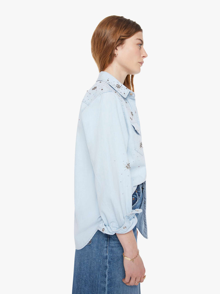 Side view of a woman in a faded light blue with faux pearl snaps down the front and beaded details with rhinestones across the shoulders and chest denim button-up with front patch pockets and a curved hem.