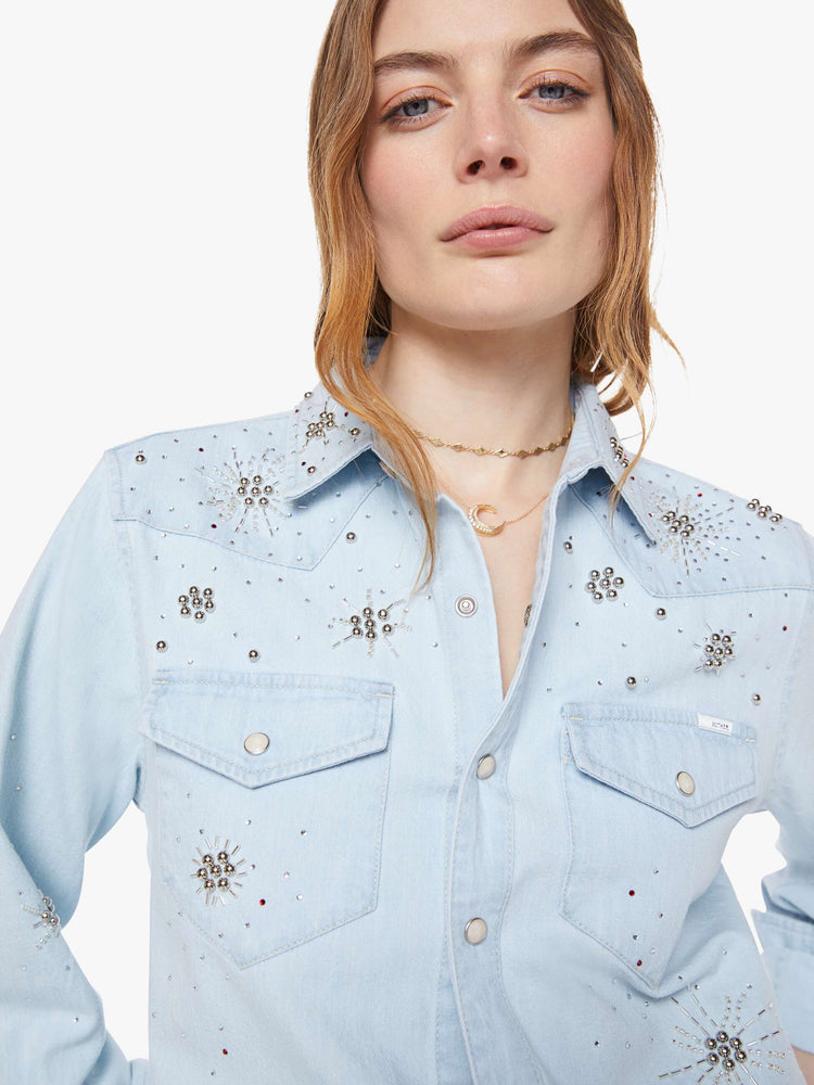 Close up view of a woman in a faded light blue with faux pearl snaps down the front and beaded details with rhinestones across the shoulders and chest denim button-up with front patch pockets and a curved hem.