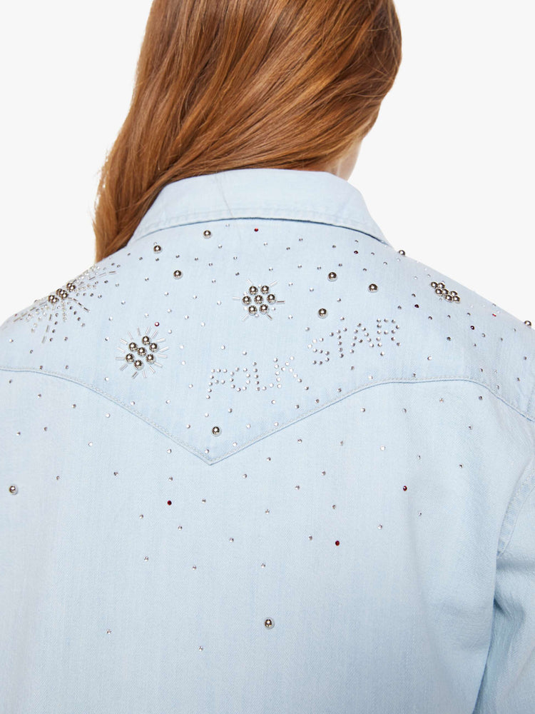 Swatch view of a woman in a faded light blue with faux pearl snaps down the front and beaded details with rhinestones across the shoulders and chest denim button-up with front patch pockets and a curved hem.