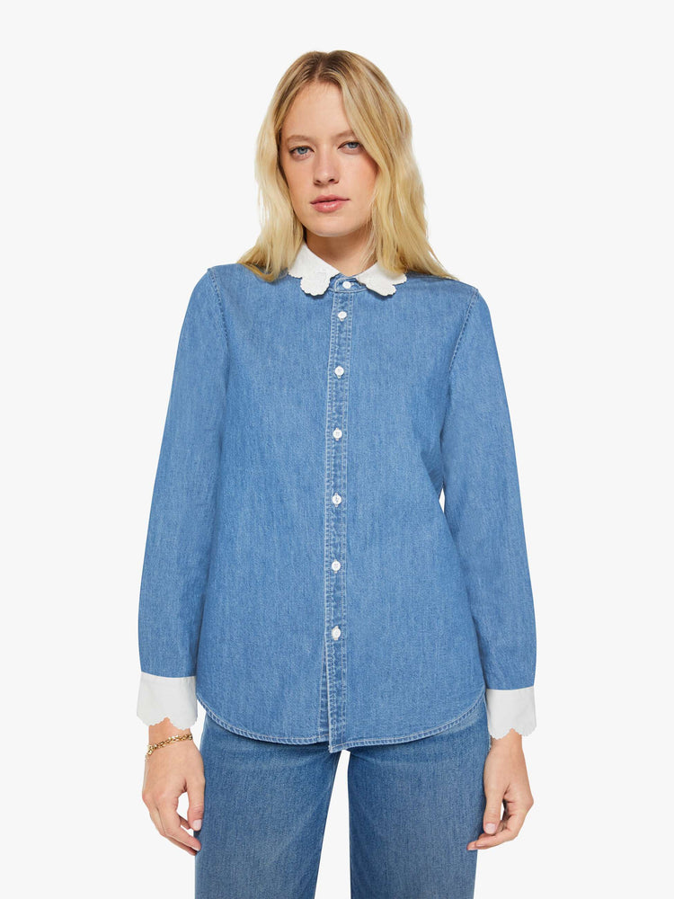 Front view of a woman in a mid-blue with fading throughout and white trim with embroidery and scalloped edges denim button-up with a curved hem and a boxy fit.