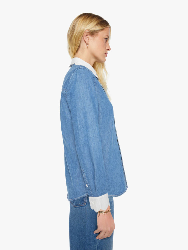 Side view of a woman in a mid-blue with fading throughout and white trim with embroidery and scalloped edges denim button-up with a curved hem and a boxy fit.