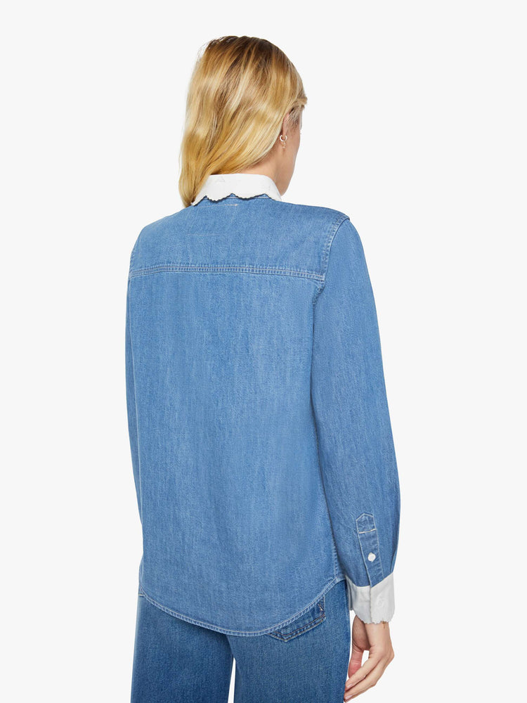 Back view of a woman in a mid-blue with fading throughout and white trim with embroidery and scalloped edges denim button-up with a curved hem and a boxy fit.