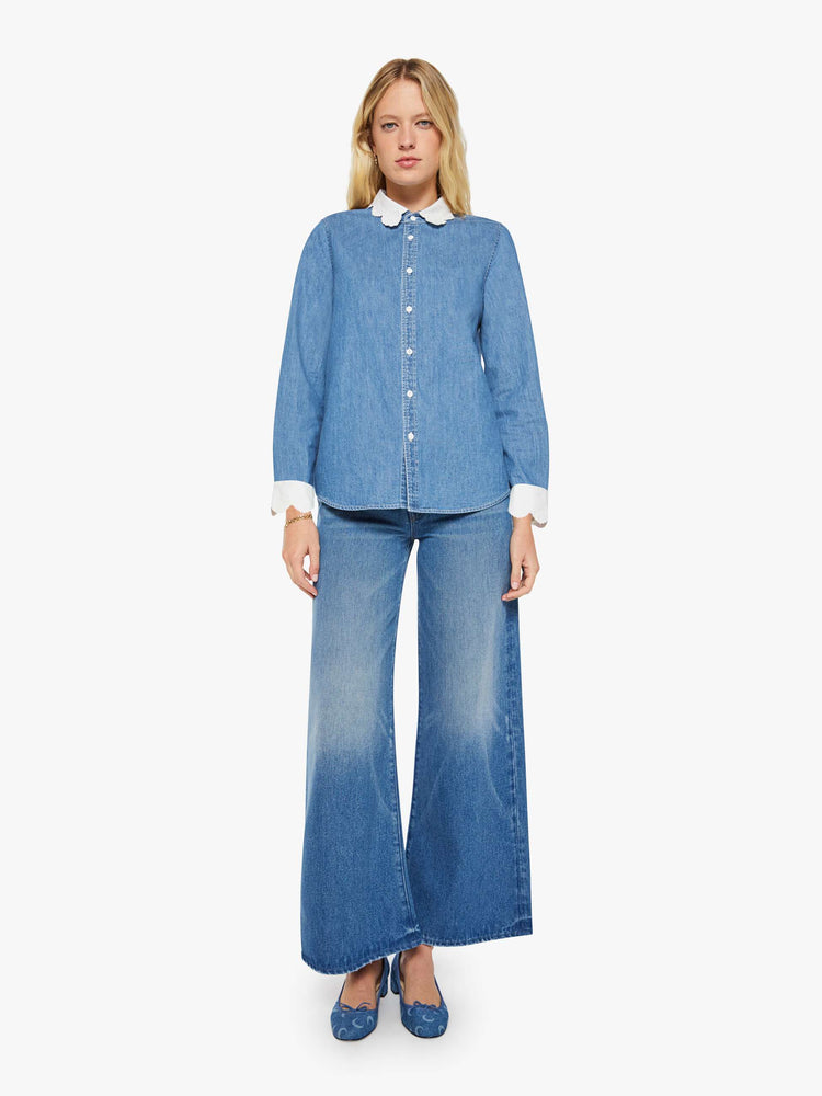 Full body view of a woman in a mid-blue with fading throughout and white trim with embroidery and scalloped edges denim button-up with a curved hem and a boxy fit.