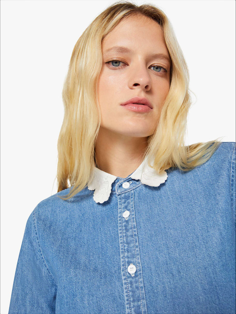 Close up view of a woman in a mid-blue with fading throughout and white trim with embroidery and scalloped edges denim button-up with a curved hem and a boxy fit.