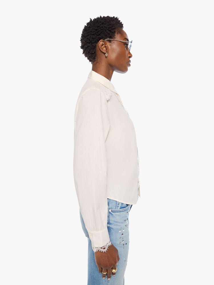Side view of a woman in top is designed in off-white button-up with a pointed collar, long balloon sleeves and a curved, cropped hem with eyelet-embroidered ruffles down the front, across the shoulders and at the wrists.