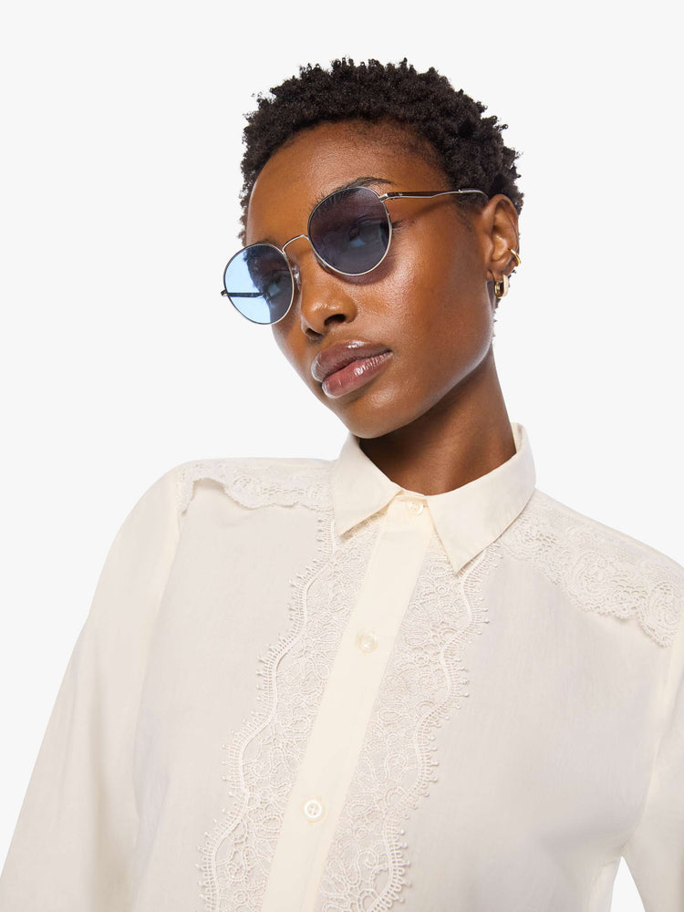 Close up view of a woman in top is designed in off-white button-up with a pointed collar, long balloon sleeves and a curved, cropped hem with eyelet-embroidered ruffles down the front, across the shoulders and at the wrists.