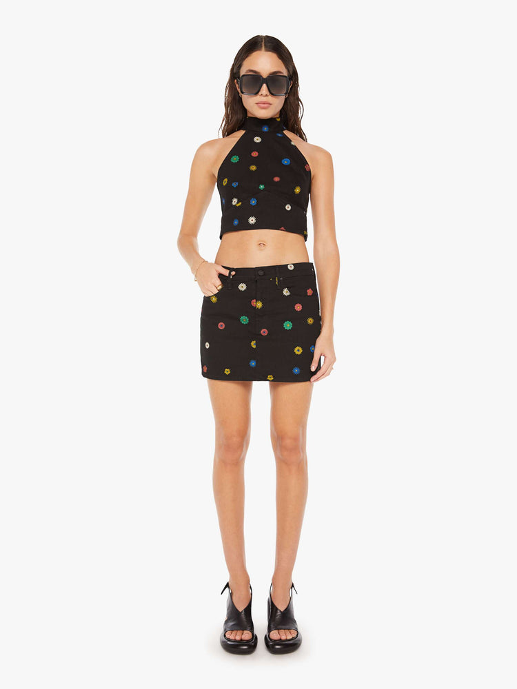 Full body view of a woman denim halter top with a mock neck, back-zip closure, darts and angled seams at the chest in black with colorful flowers printed throughout.