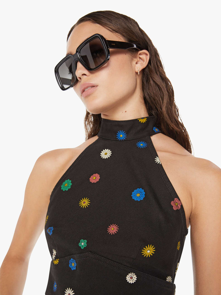 Close up view of a woman denim halter top with a mock neck, back-zip closure, darts and angled seams at the chest in black with colorful flowers printed throughout.
