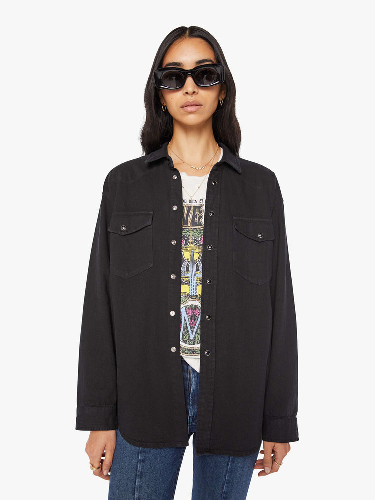 Front view of woman in a black oversized denim button-up with drop shoulders, front patch pockets, a Western-inspired yoke and a curved hem.