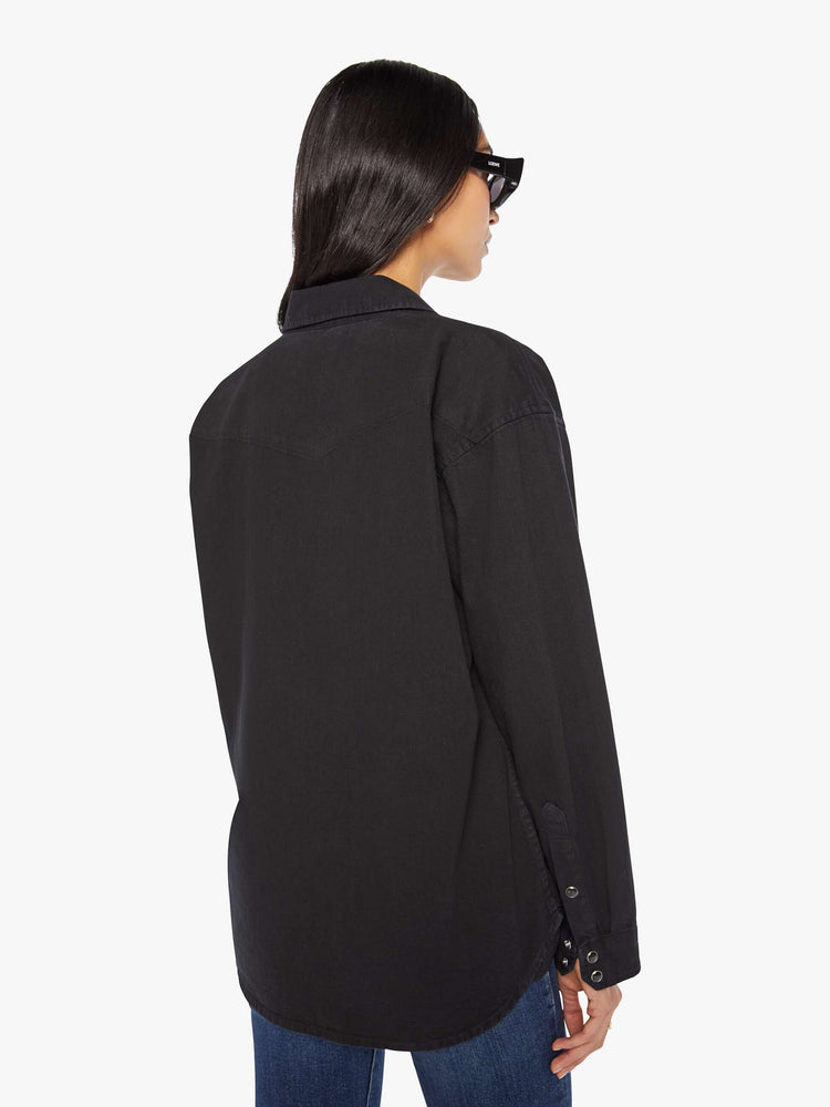 Back view of woman in a black oversized denim button-up with drop shoulders, front patch pockets, a Western-inspired yoke and a curved hem.