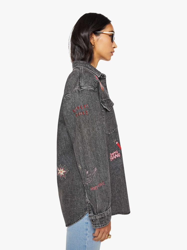Side view of a woman in a faded dark grey with embroidered graphics throughout  oversized denim button-up with drop shoulders, front patch pockets, a Western-inspired yoke and a curved hem. 