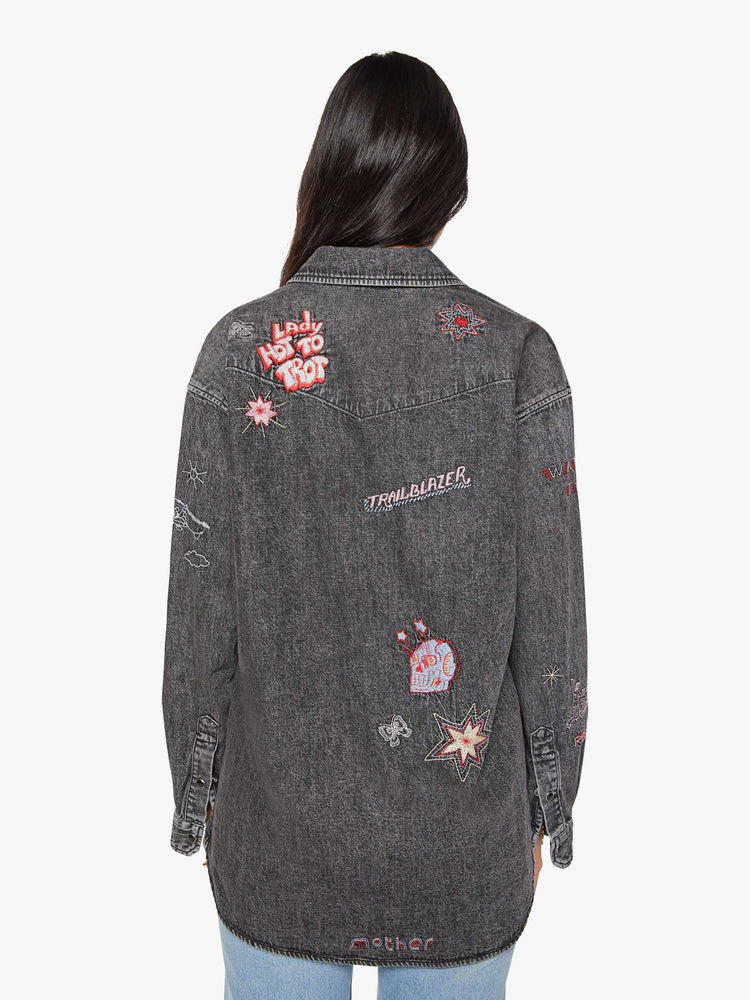 Back view of a woman in a faded dark grey with embroidered graphics throughout  oversized denim button-up with drop shoulders, front patch pockets, a Western-inspired yoke and a curved hem. 