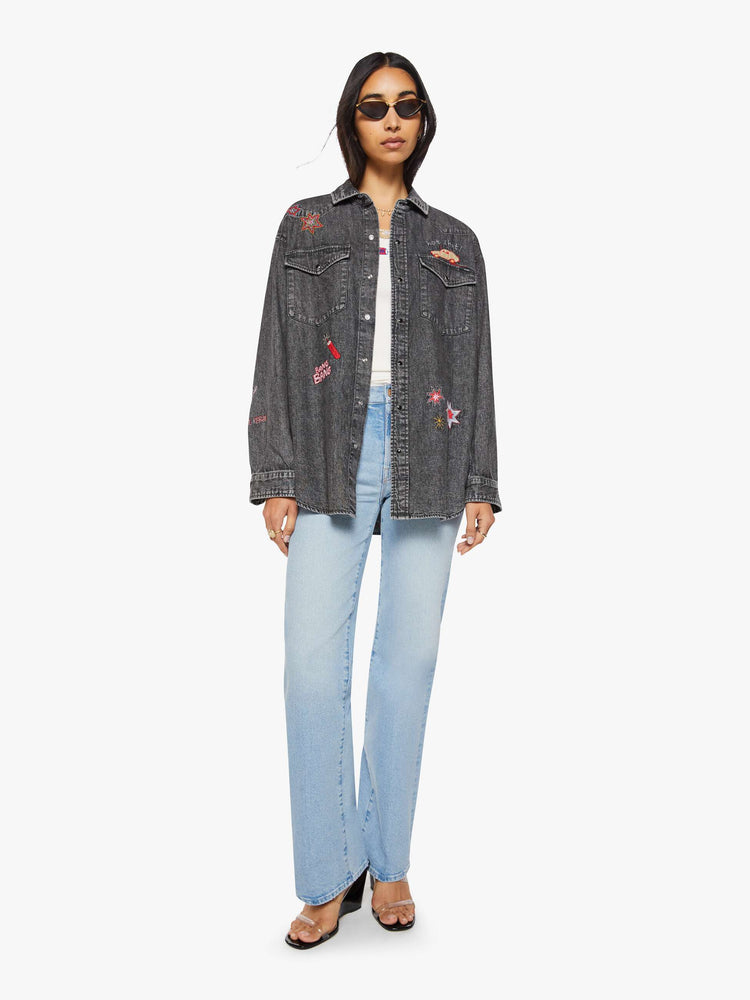 Full body view of a woman in a faded dark grey with embroidered graphics throughout  oversized denim button-up with drop shoulders, front patch pockets, a Western-inspired yoke and a curved hem. 