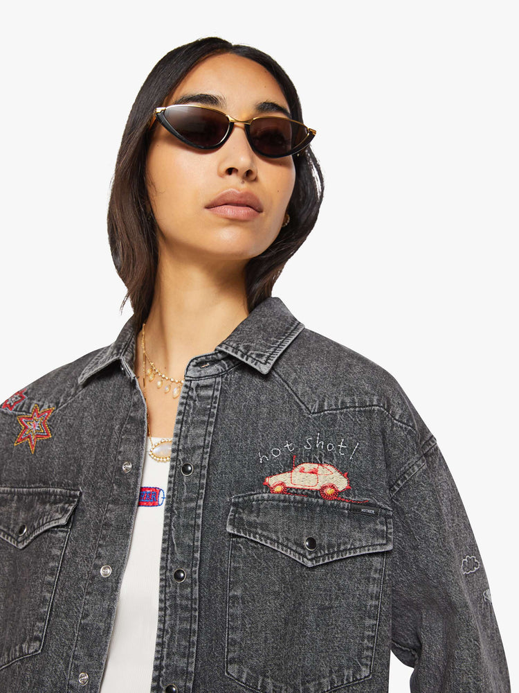 Close up view of a woman in a faded dark grey with embroidered graphics throughout  oversized denim button-up with drop shoulders, front patch pockets, a Western-inspired yoke and a curved hem. 