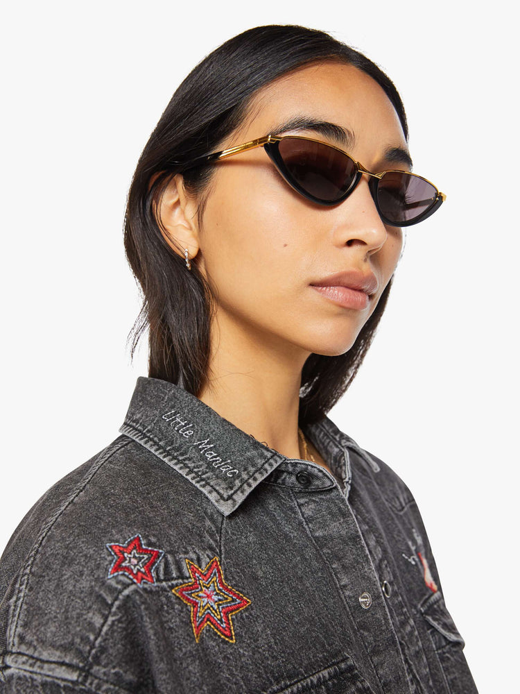 Close up view of a woman in a faded dark grey with embroidered graphics throughout  oversized denim button-up with drop shoulders, front patch pockets, a Western-inspired yoke and a curved hem. 