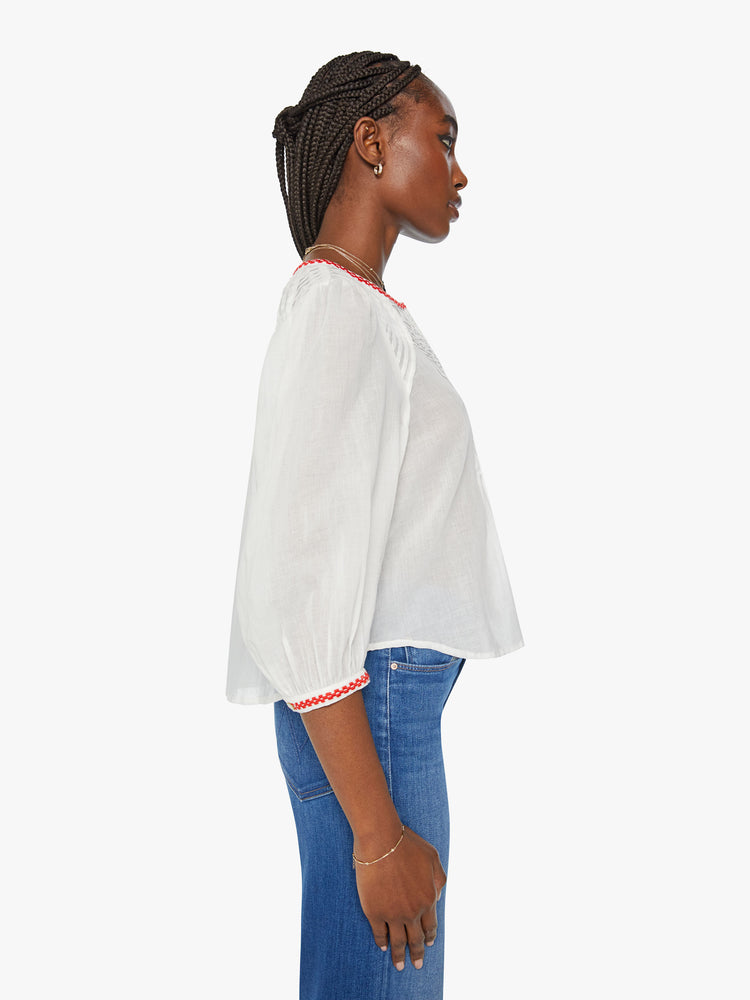 Side view of a woman white blouse blouse with a crewneck, 3/4-length balloon sleeves, pleated details, buttons down the front and a slightly shrunken fit in white with red trim around the neck and sleeves.
