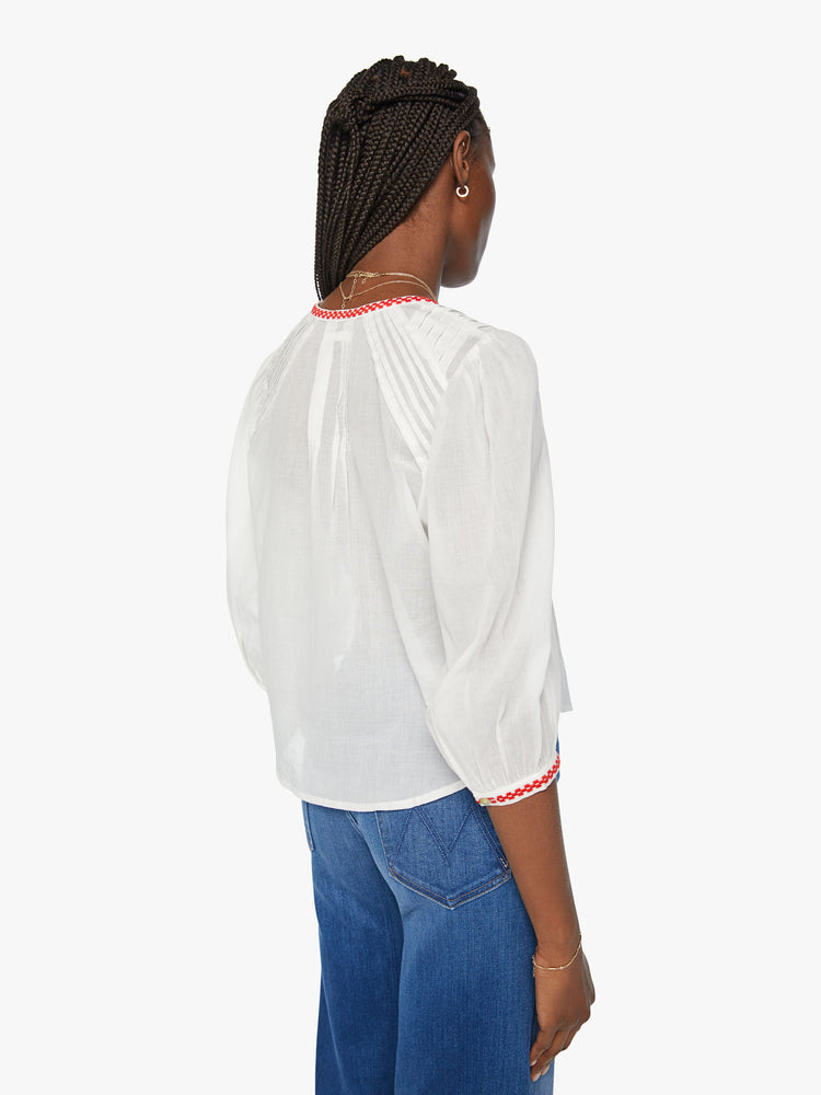 Back view of a woman white blouse blouse with a crewneck, 3/4-length balloon sleeves, pleated details, buttons down the front and a slightly shrunken fit in white with red trim around the neck and sleeves.