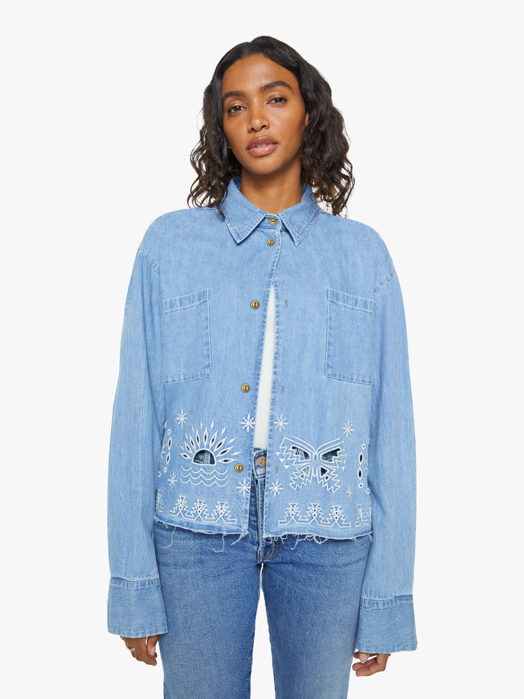 Front view of a woman in a denim button-up with drop shoulders, an oversized patch pocket and a cropped, frayed hem in light blue with embroidered details in white.