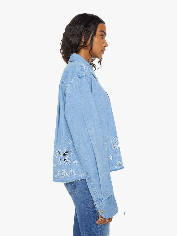 Side view of a woman in a denim button-up with drop shoulders, an oversized patch pocket and a cropped, frayed hem in light blue with embroidered details in white.