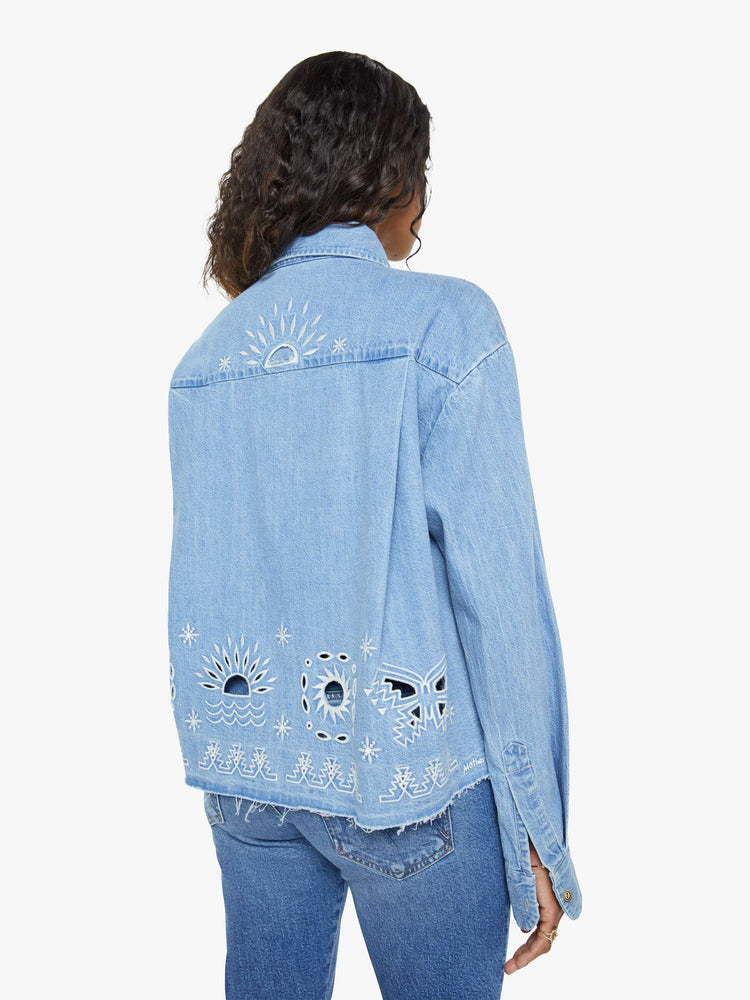 Back view of a woman in a denim button-up with drop shoulders, an oversized patch pocket and a cropped, frayed hem in light blue with embroidered details in white.