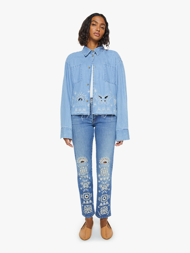 Front view of a woman in a denim button-up with drop shoulders, an oversized patch pocket and a cropped, frayed hem in light blue with embroidered details in white.