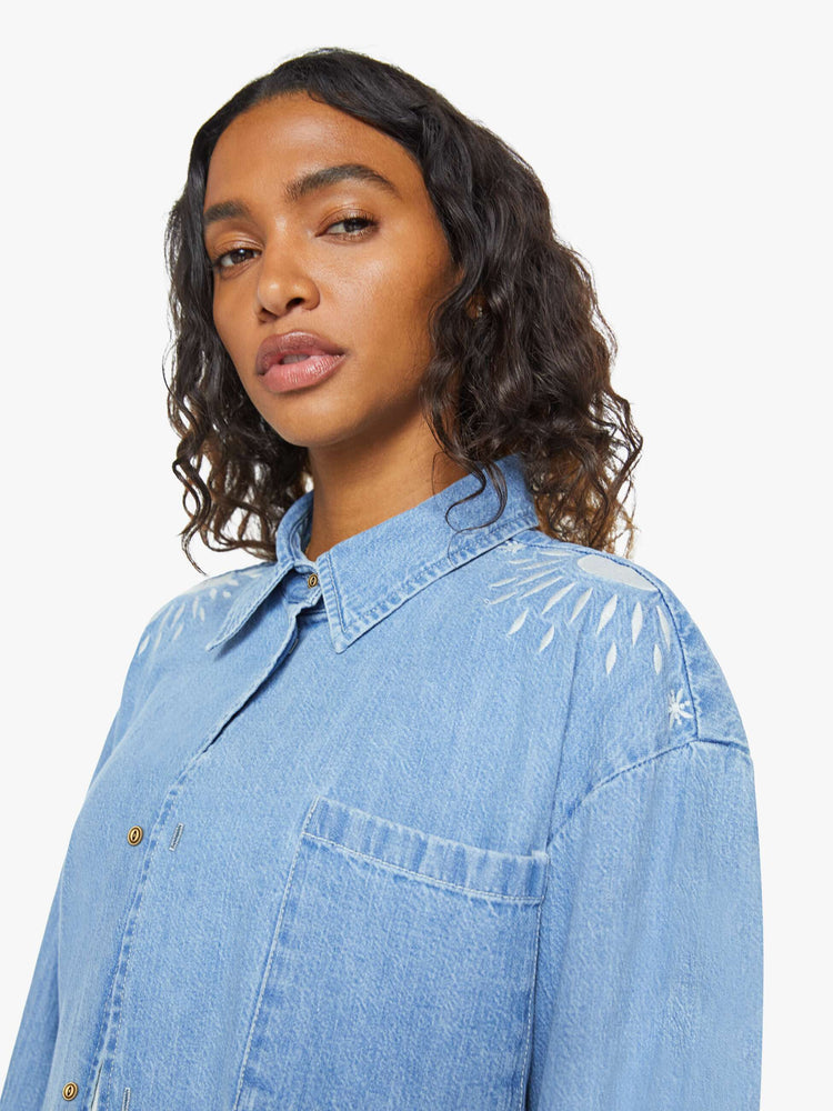 Close up view of a woman in a denim button-up with drop shoulders, an oversized patch pocket and a cropped, frayed hem in light blue with embroidered details in white.