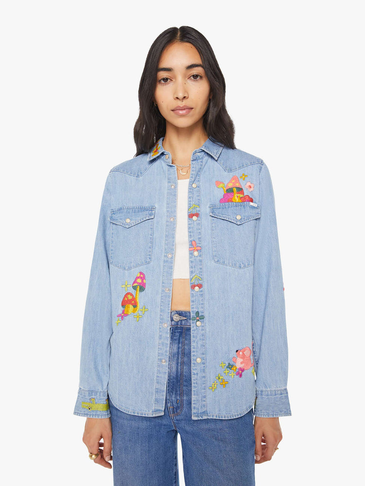 Front view of a woman in a light blue denim button-up with front patch pockets, a curved hem and slightly oversized fit with embroidery mushrooms and flowers.
