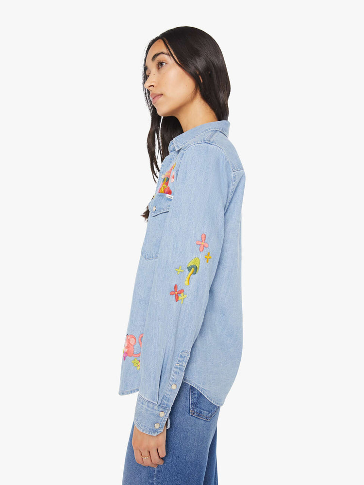 Side view of a woman in a light blue denim button-up with front patch pockets, a curved hem and slightly oversized fit with embroidery mushrooms and flowers.