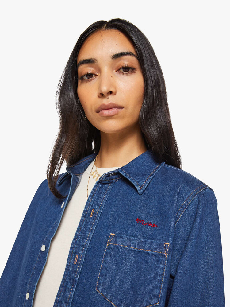 Close up view of a woman in a dark blue  cropped button-up shirt with a patch pocket and MOTHER's name embroidered in red on the chest. 