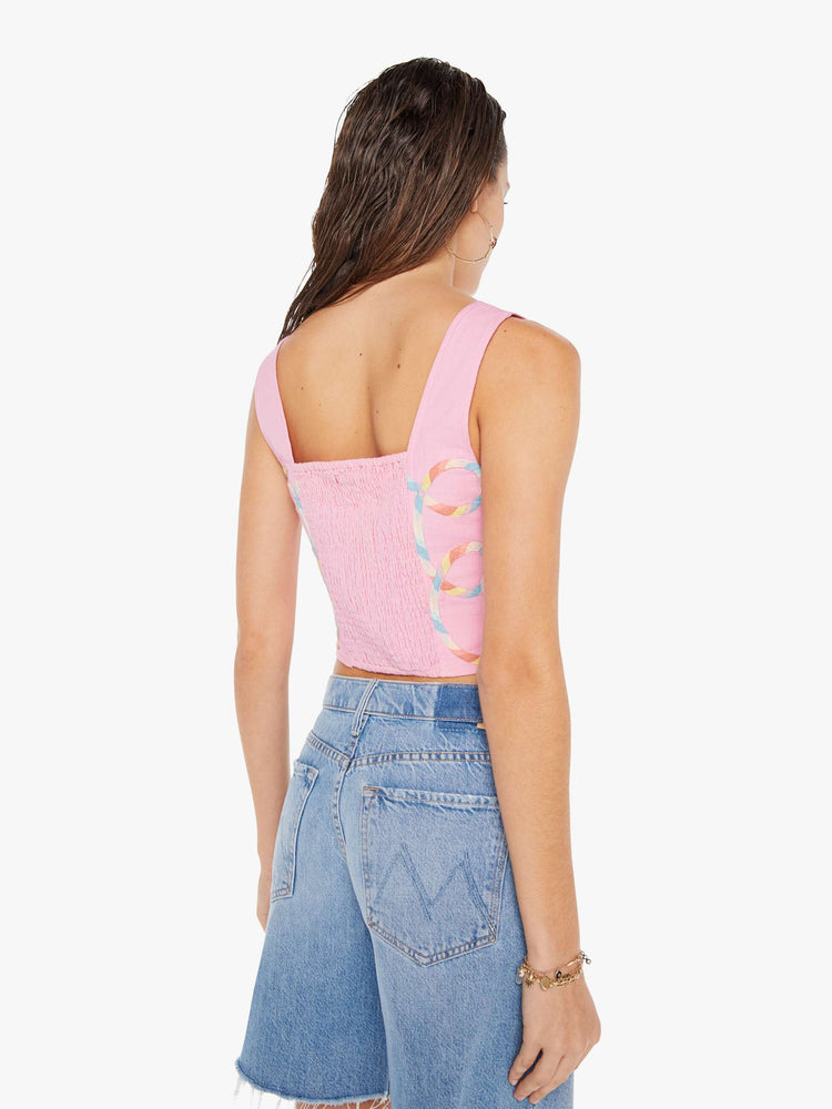 Back view of a woman wearing a pink bustier top featuring multi color ribbon embroidered throughout, paired with a light blue wash denim short.