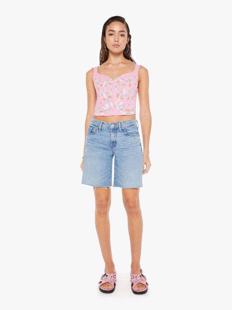 Front full body view of a woman wearing a pink bustier top featuring multi color ribbon embroidered throughout, paired with a light blue wash denim short.