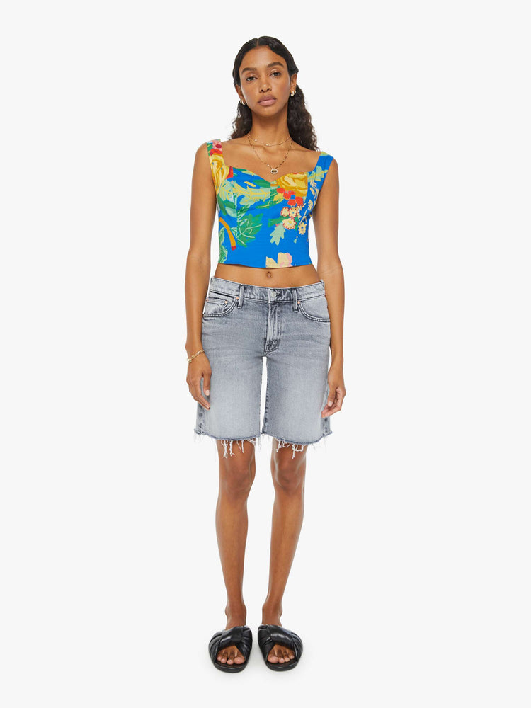 Full front view of a woman in a blue sleeveless corset with an oversized tropical floral print, a sweetheart neckline, seamed details, a smocked, elastic back panel and a cropped, narrow fit. Paired with grey shorts and black sandals.